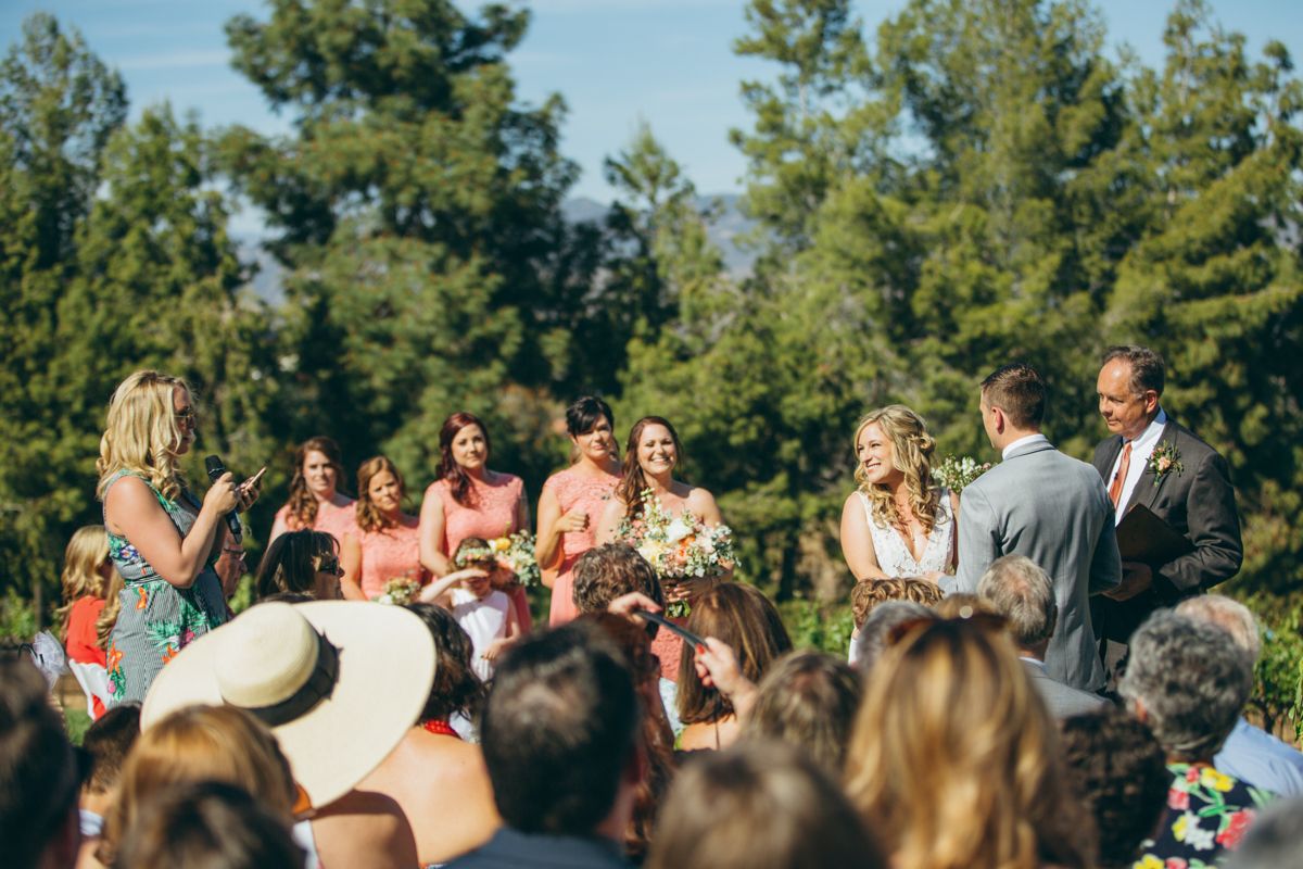 Outdoor wedding ceremony with bridesmaids in pink dresses surrounded by lush green trees.
