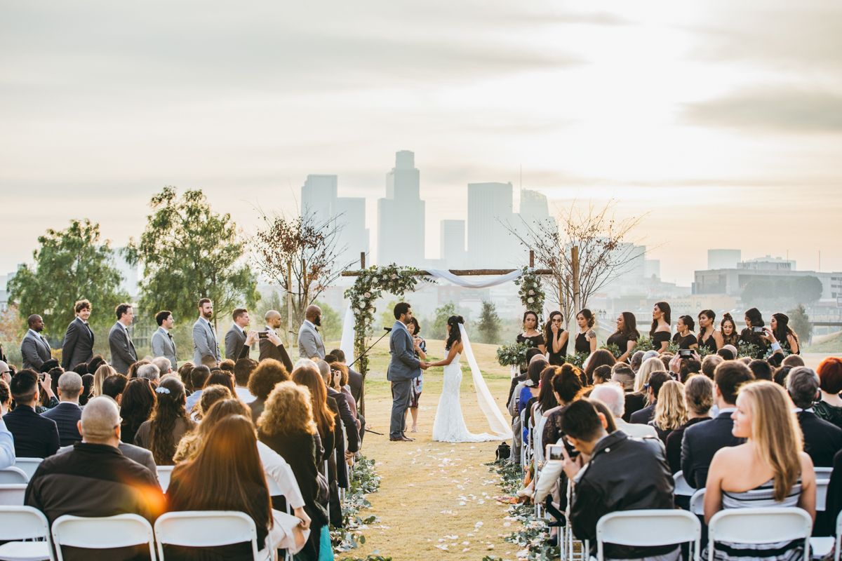 Outdoor wedding ceremony with couple at floral arch altar, guests seated in white chairs, city skyline backdrop at sunset.