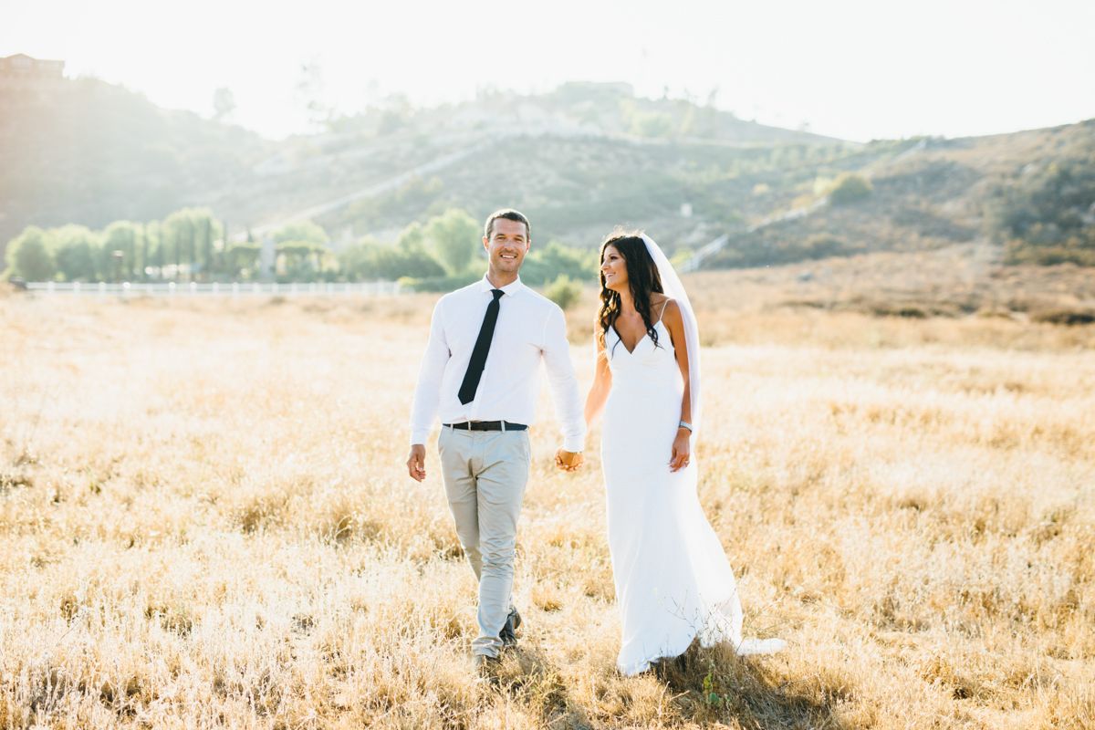 A bride and groom hold hands walking through a golden sunlit field with rolling hills behind them.