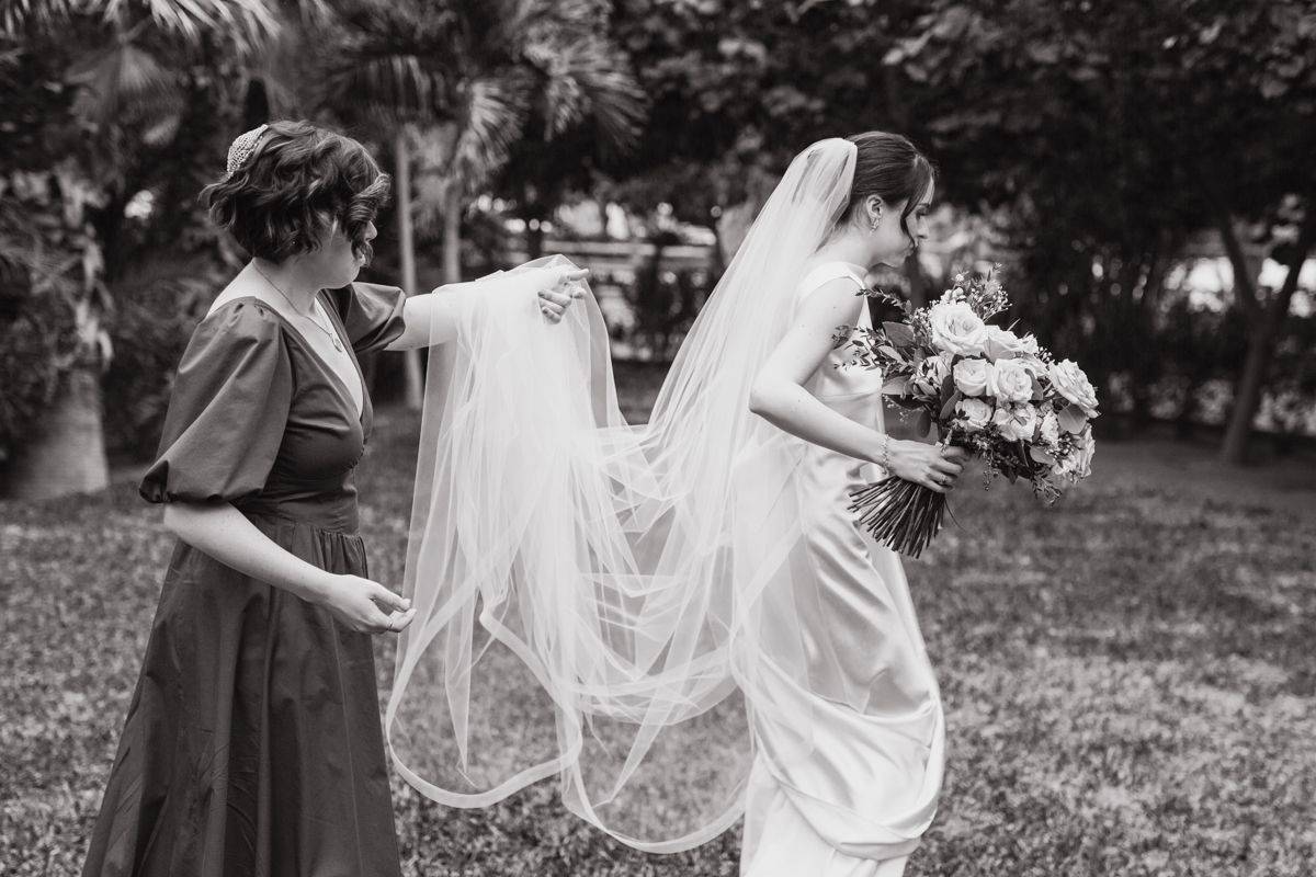 Bridesmaid helps arrange bride's veil outdoors in black and white wedding photo.