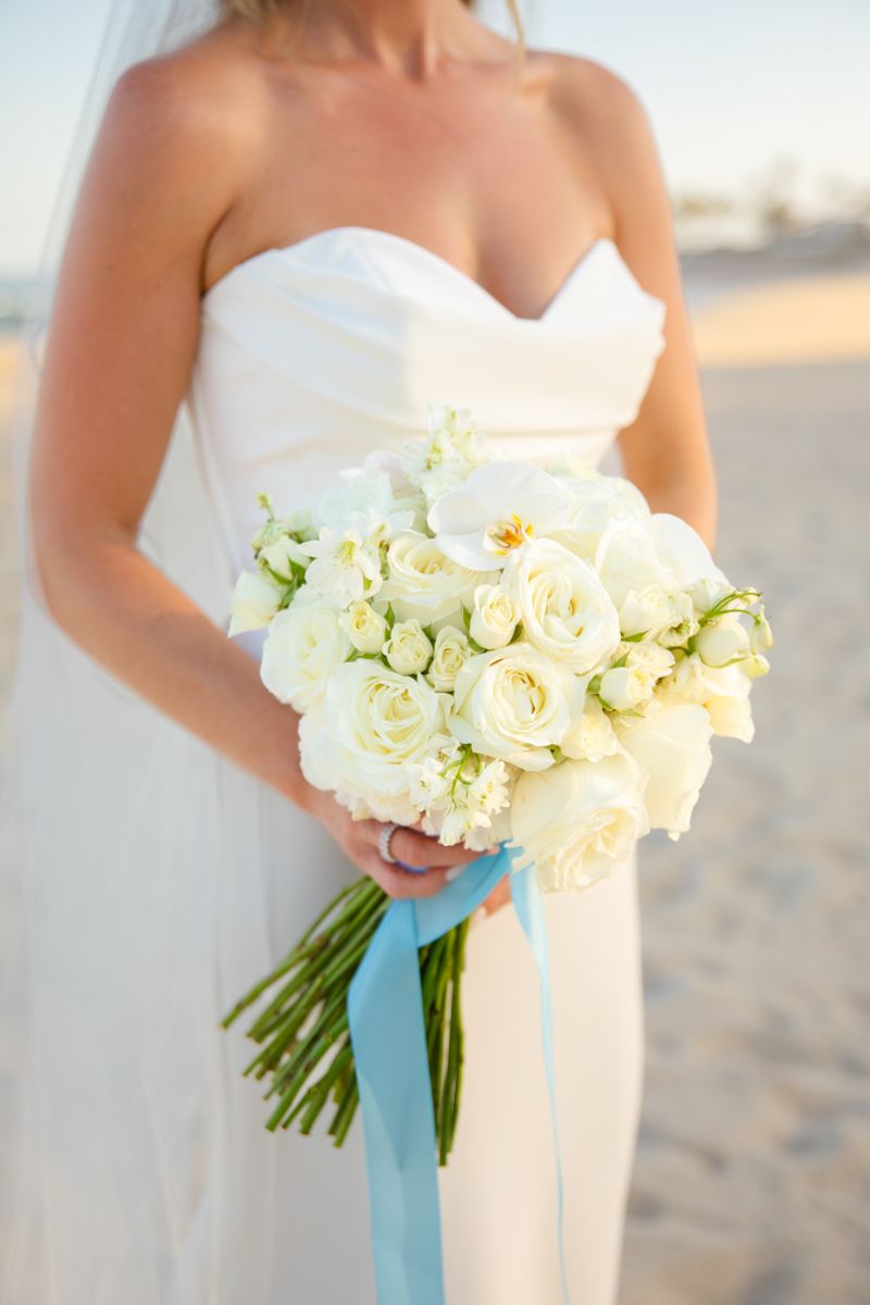Close-up of bride holding elegant white rose bouquet tied with a light blue ribbon.