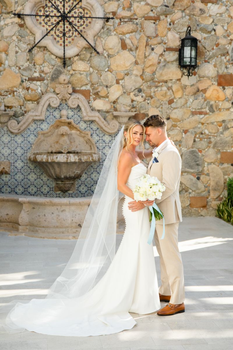 Bride and groom posing together near stone wall fountain, bride in long veil.