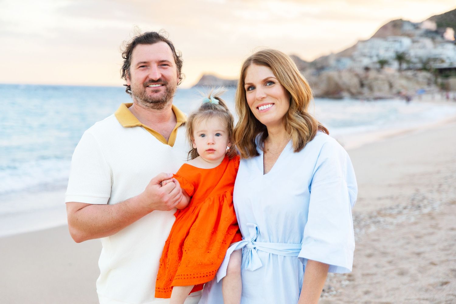 Young family of three posing on beach at sunset, baby girl in orange dress.