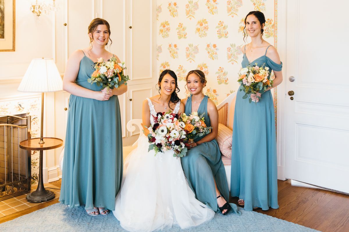 Bride seated with three bridesmaids in teal gowns holding bouquets in a elegantly decorated room.