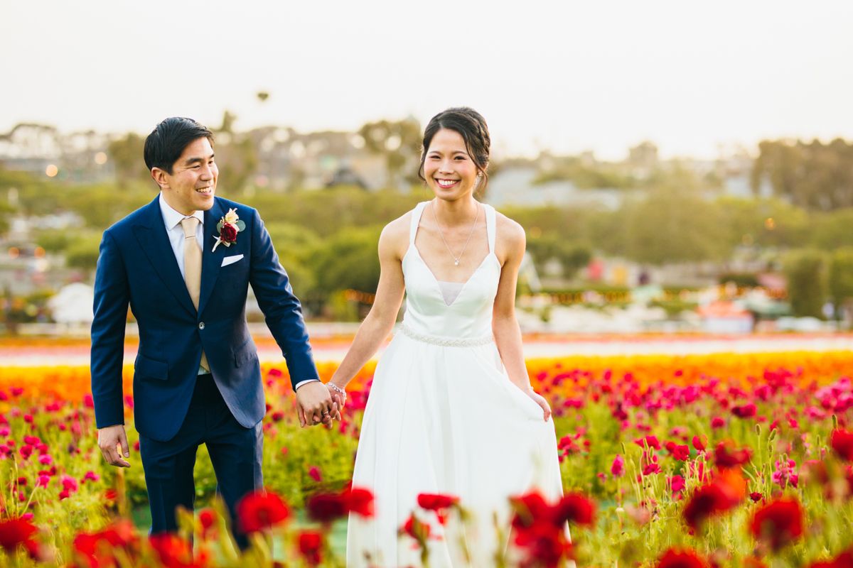 Happy wedding couple holding hands walking through a vibrant red and pink flower field.