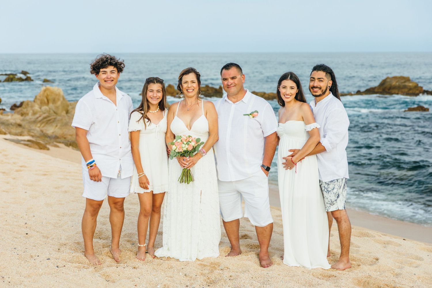Family group photo on sandy beach with ocean backdrop, dressed in white for a cabo wedding celebration.