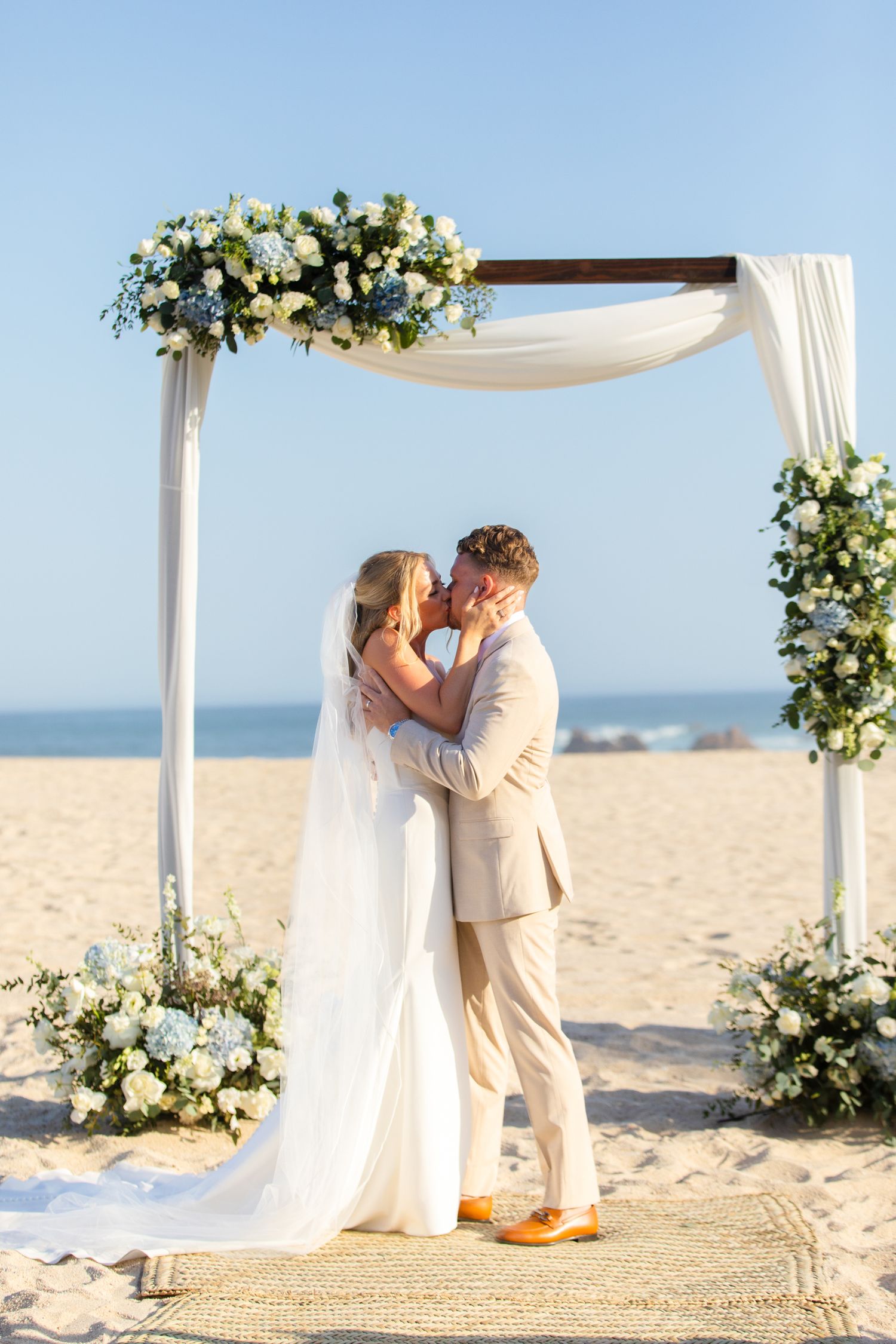 Couple sharing first kiss under floral wedding arch on sandy beach with blue hydrangeas and white draping.