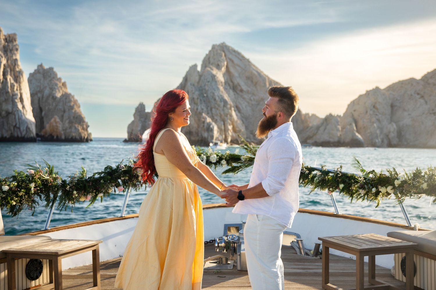 Couple holds hands on yacht deck with rocky ocean backdrop at sunset, woman in yellow dress, man in white suit.