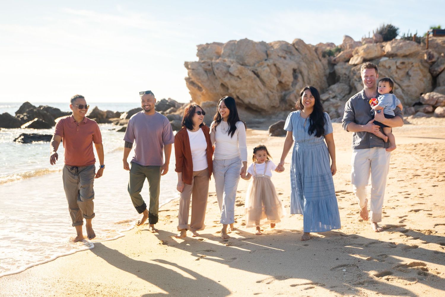 Large family group walking together on a sunny beach in Cabo San Lucas with rocky cliffs, enjoying a seaside outing.