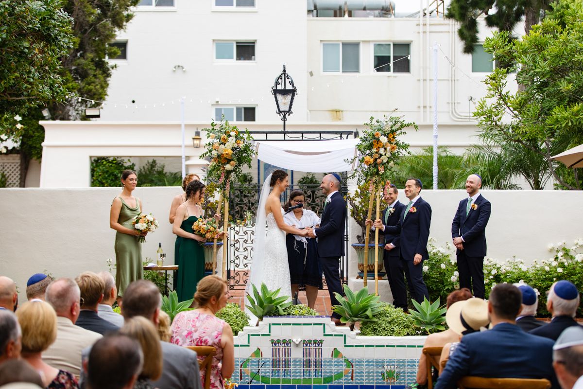 Outdoor Jewish wedding ceremony with wedding party at floral chuppah, guests seated on brick patio.