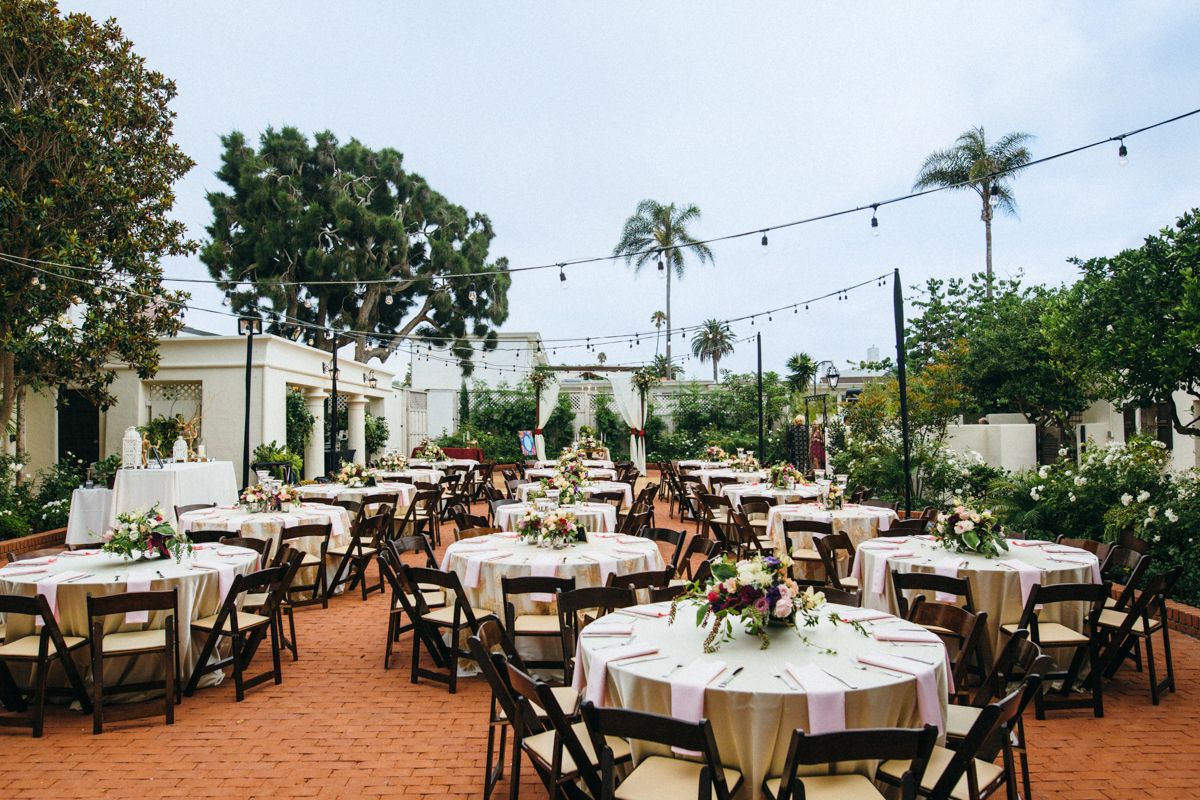 Outdoor wedding reception with round tables, dark folding chairs, floral centerpieces, string lights, and lush greenery.