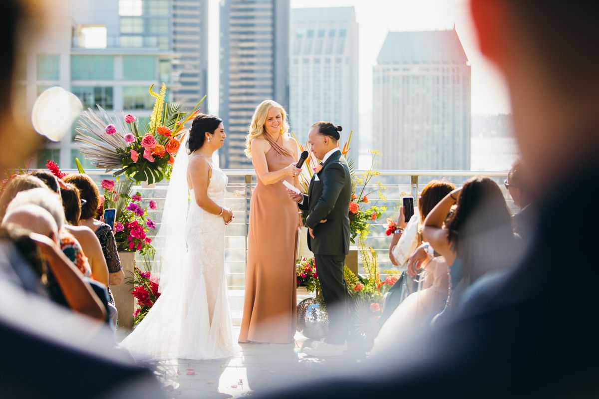 Rooftop wedding ceremony with officiant speaking as bride and groom stand before colorful floral arrangements.