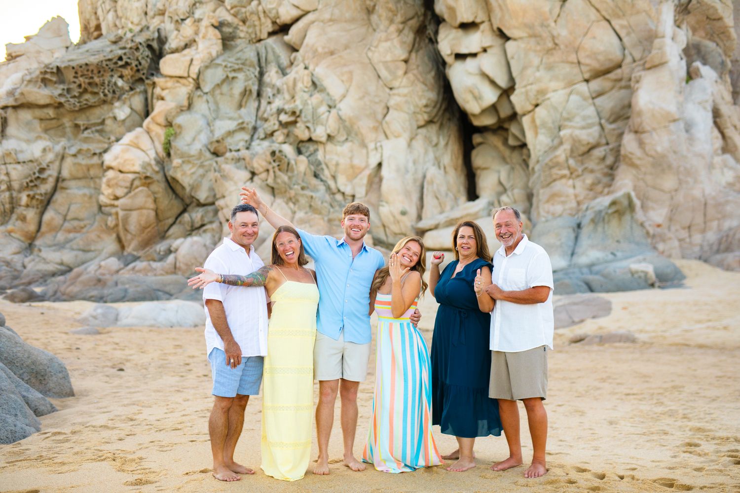 Six people smiling together on a sandy beach with large rocky formations in the background on a sunny day.