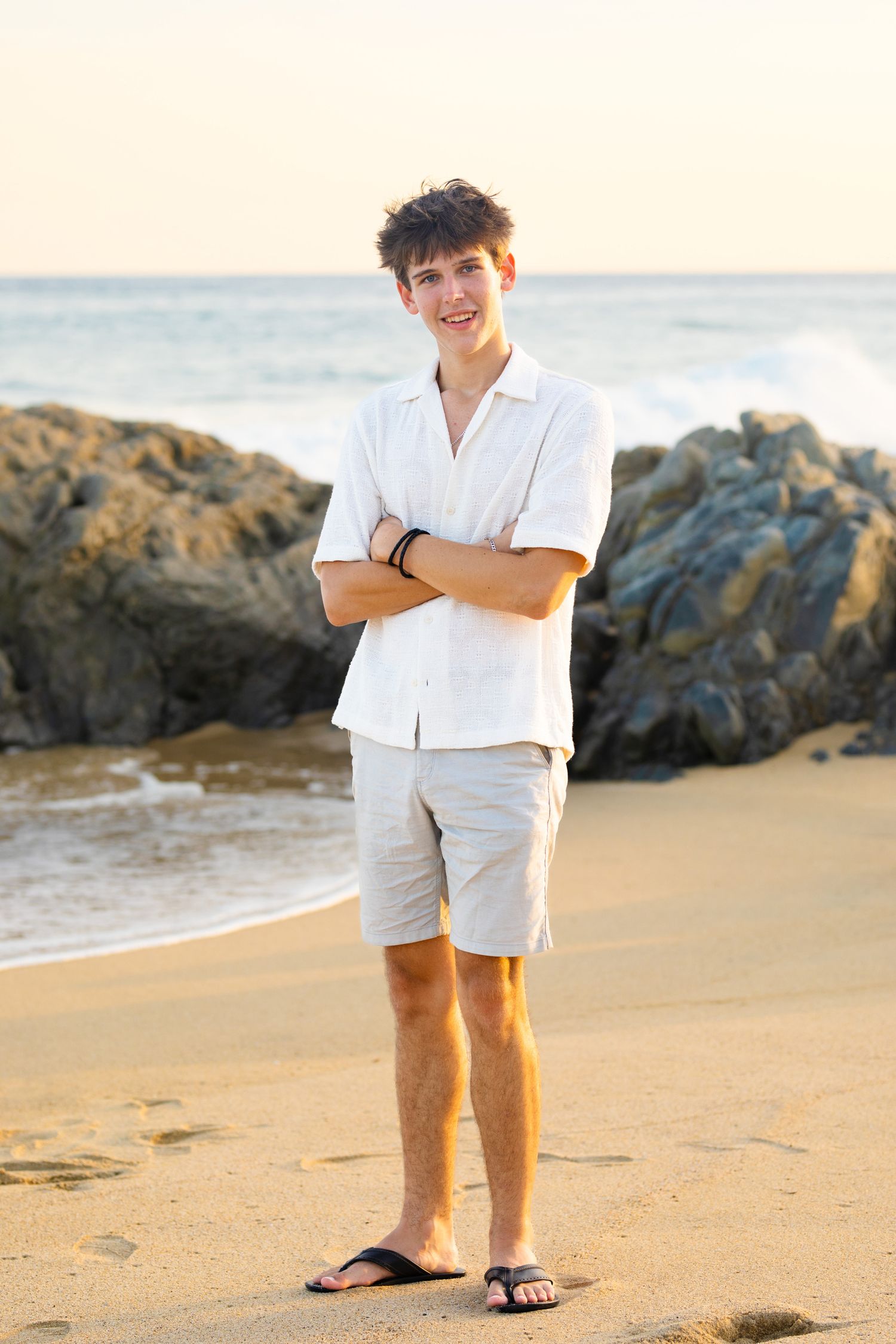 Smiling teen boy in white shirt and khaki shorts standing arms crossed on sandy beach.