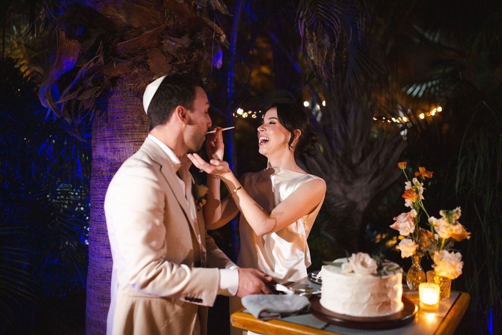 Bride feeding wedding cake to groom wearing a kippah outdoors at night with floral arrangements nearby.