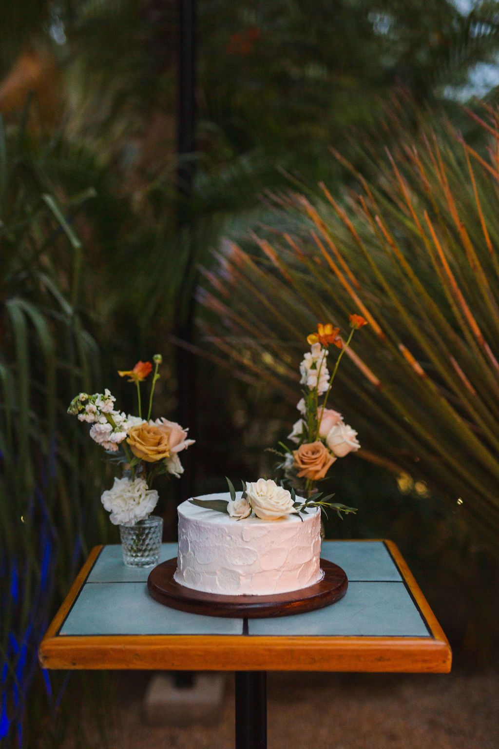 Single-tier white wedding cake decorated with peach and white fresh flowers on a wooden cake stand outdoors.