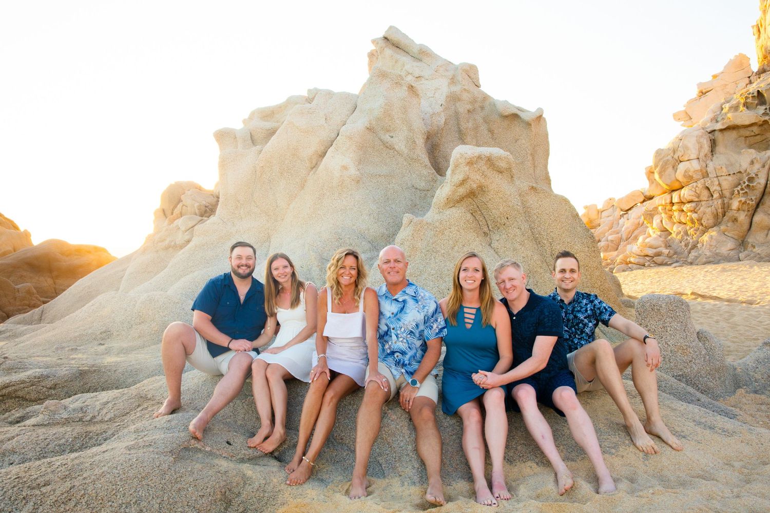 Family of seven smiling together on rocky beach boulders at golden sunset in Cabo San Lucas.