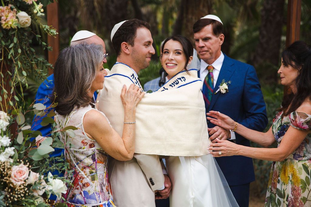 A bride and groom share a warm embrace with family under a tallit during their outdoor Jewish wedding ceremony.