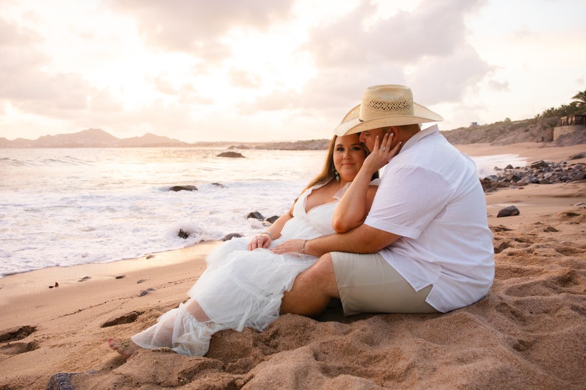 Couple sitting together on sandy beach at sunset, woman in white dress leaning against man in straw hat.