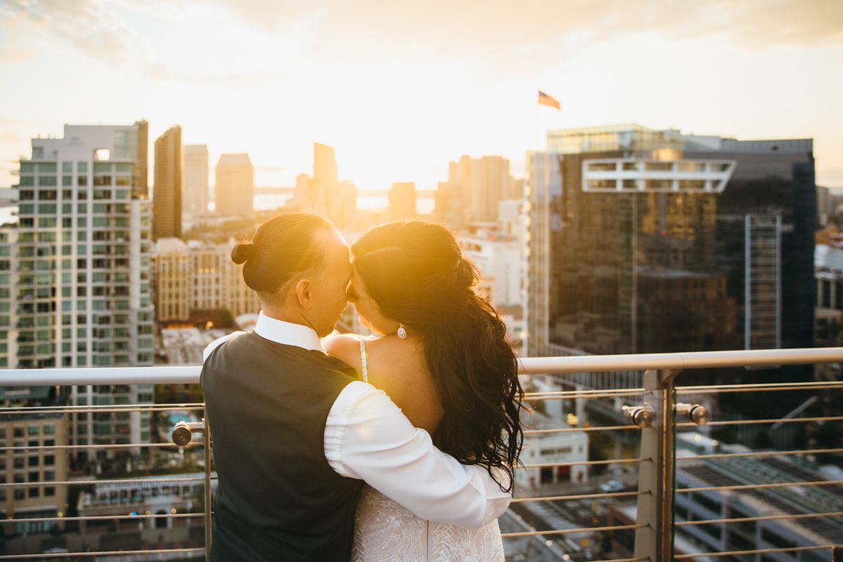 Couple shares romantic kiss on rooftop at golden sunset with city skyline in background.