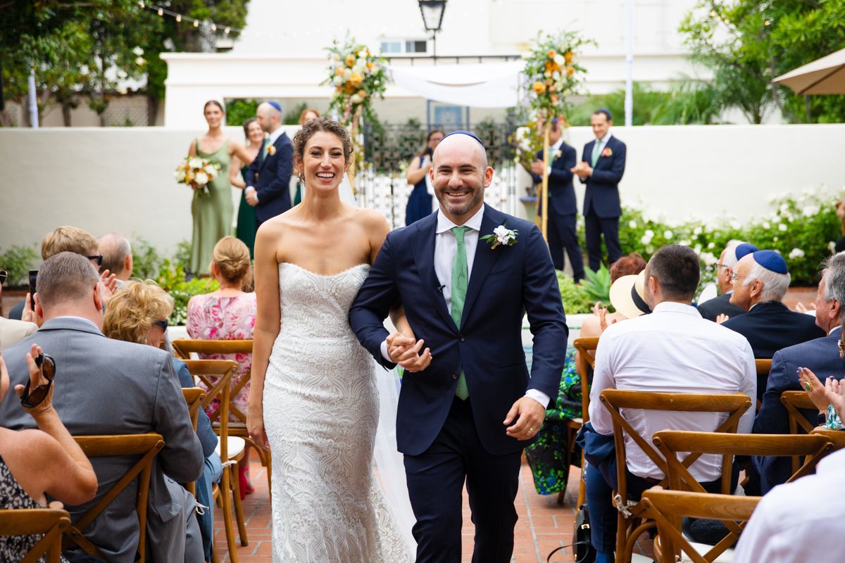 Newlyweds walk joyfully down aisle after ceremony as guests applaud in outdoor garden venue.