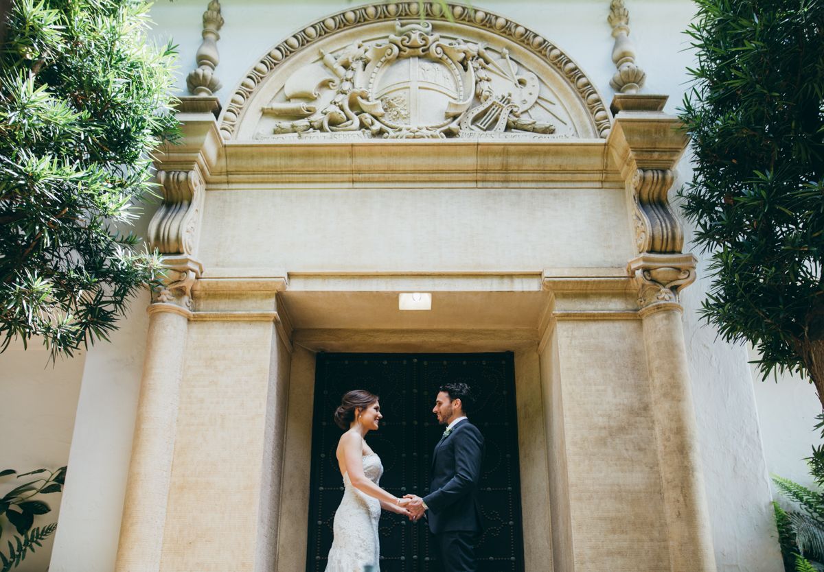 Bride and groom hold hands facing each other before an ornate church doorway with classical stone carvings.