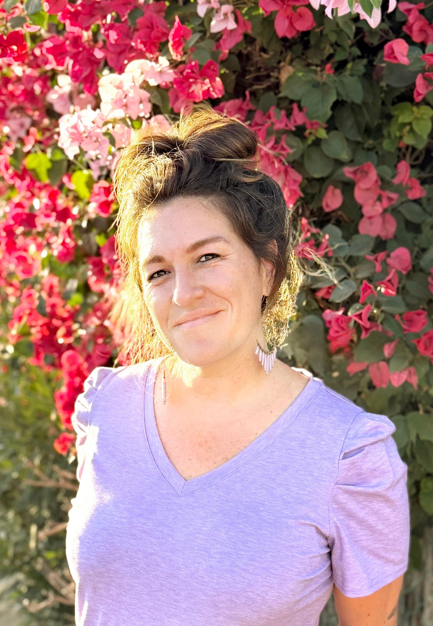 Smiling woman with hair up in bun wearing lavender top, standing before vibrant pink bougainvillea flowers.