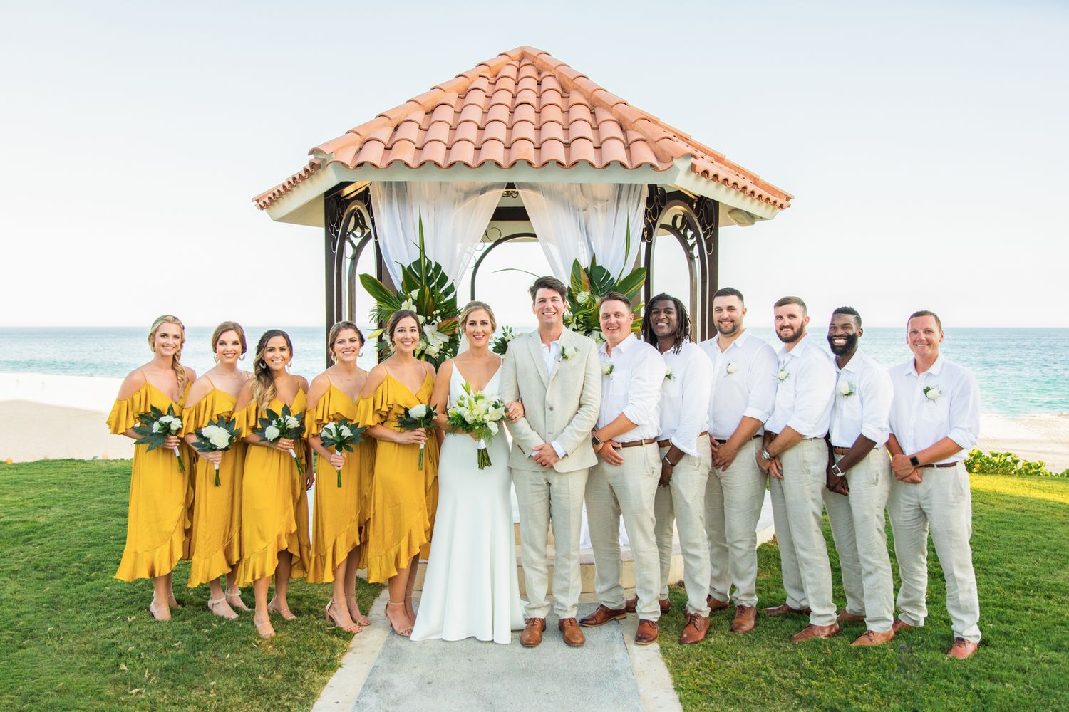 Wedding party poses by oceanside gazebo, bridesmaids in yellow dresses, groomsmen in tan suits, bride and groom at center.