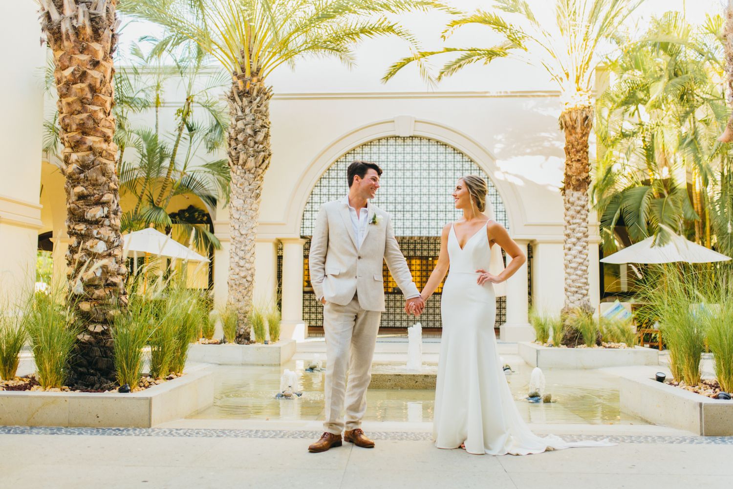 Bride and groom hold hands in a sunlit courtyard with palm trees and white umbrellas at an elegant outdoor wedding venue.
