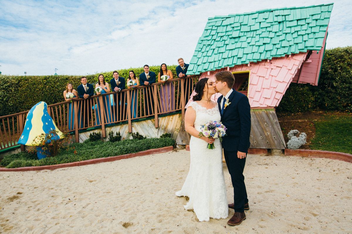 Bride and groom kiss on sandy ground with wedding party celebrating on balcony above.