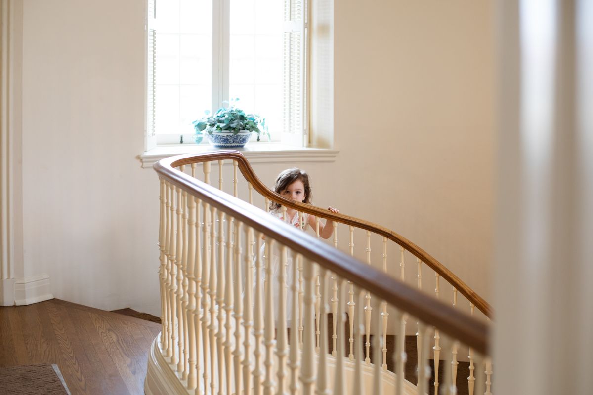 Young flower girl peeks through staircase railing in bright white wedding venue.