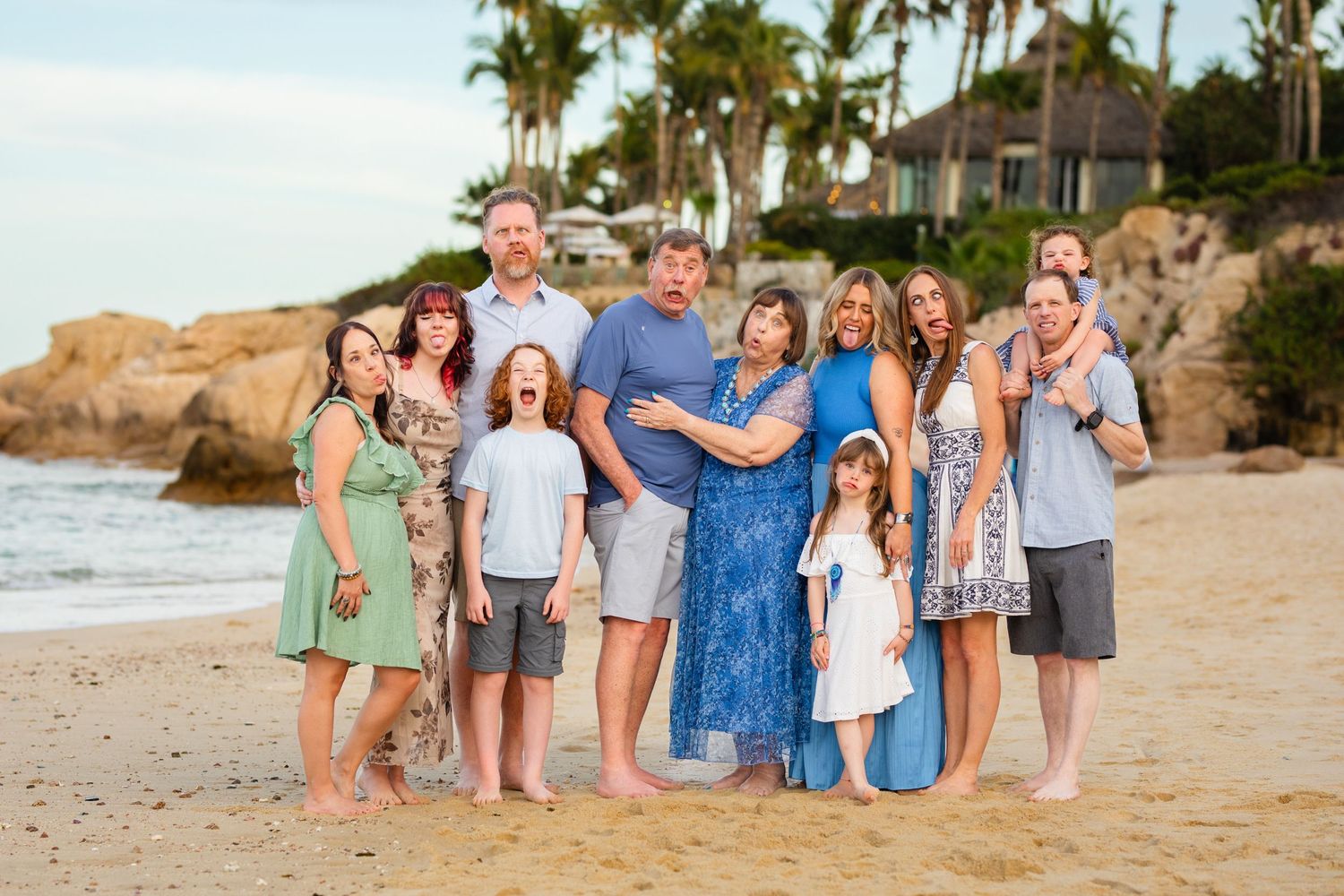 Large family group posing together on a sandy beach in cabo at sunset with palm trees and ocean in the background.