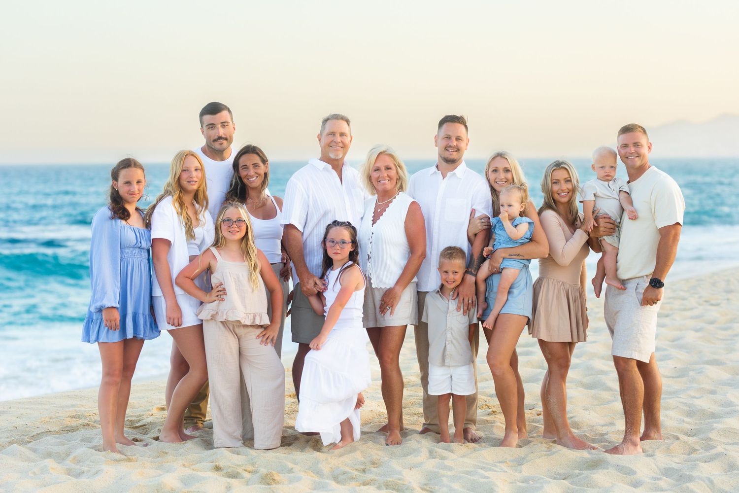 Large family group portrait on a sunny beach in cabo,  adults and children smiling in coordinated outfits with ocean waves behind.