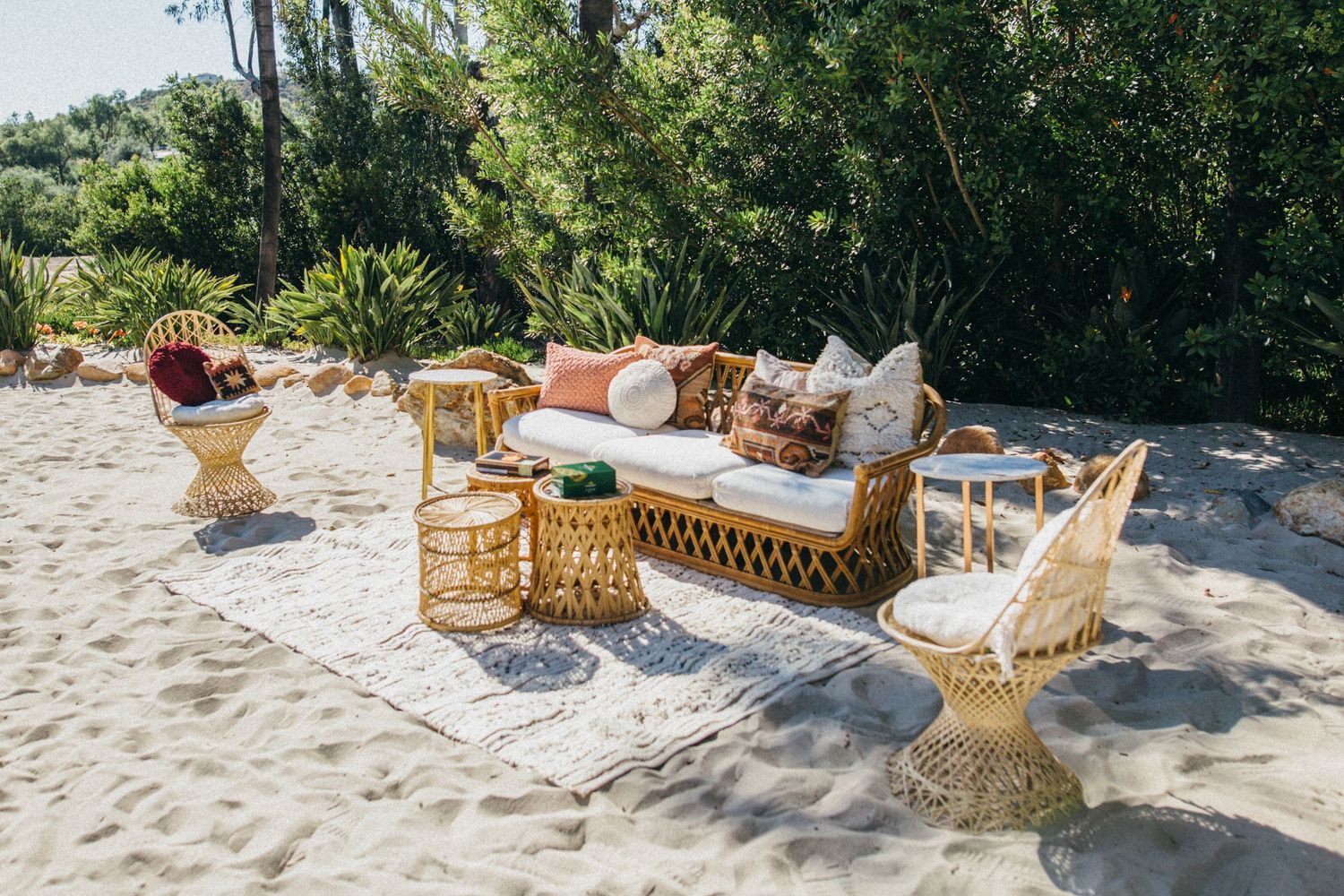 Bohemian rattan furniture set with white cushions and colorful pillows arranged on a sandy beach with lush green trees.