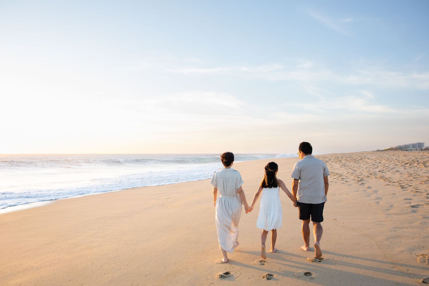 Family of three walking hand in hand along a serene beach at dusk.