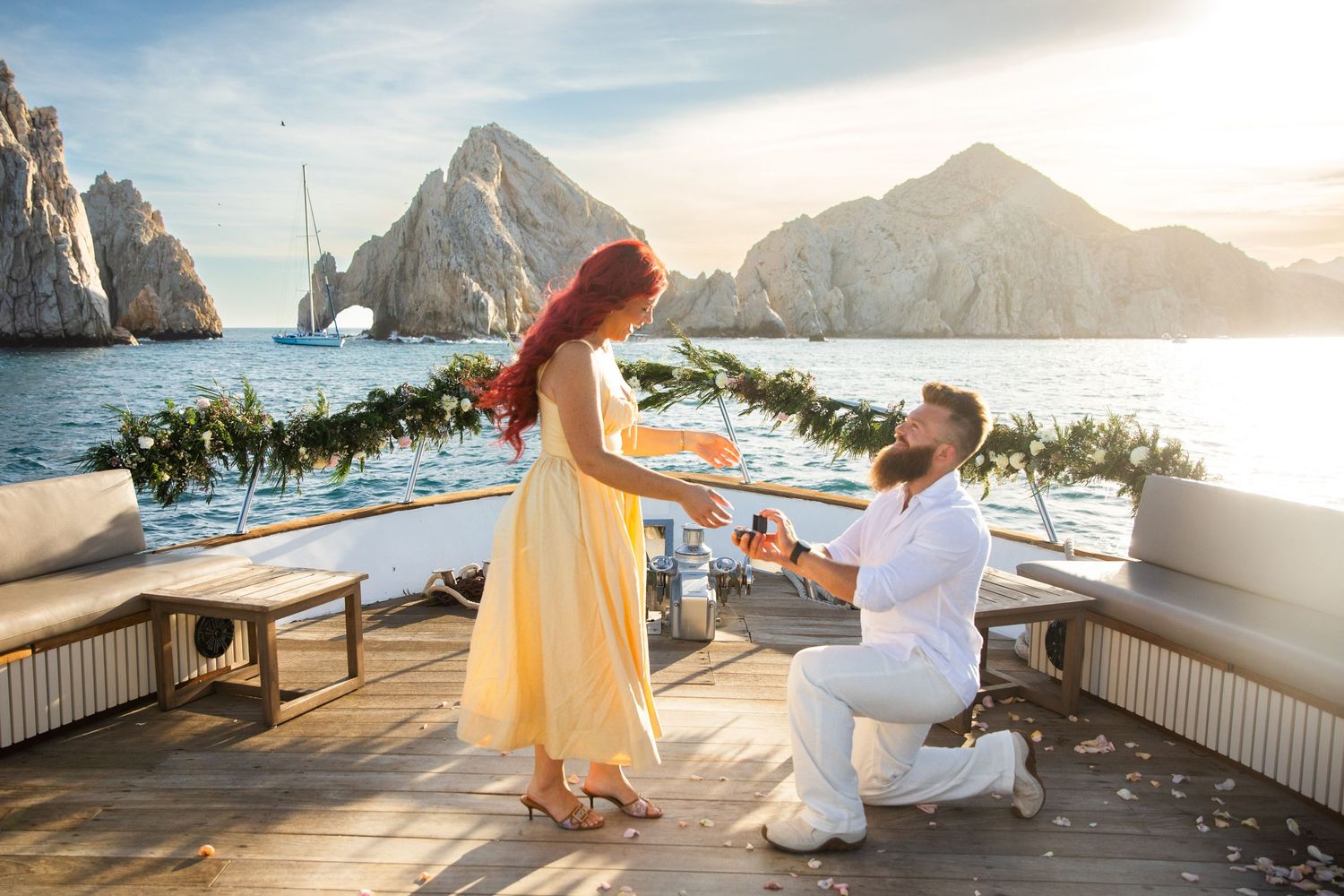 Man proposes to woman on yacht deck with scenic rocky ocean backdrop at sunset in Cabo San Lucas.
