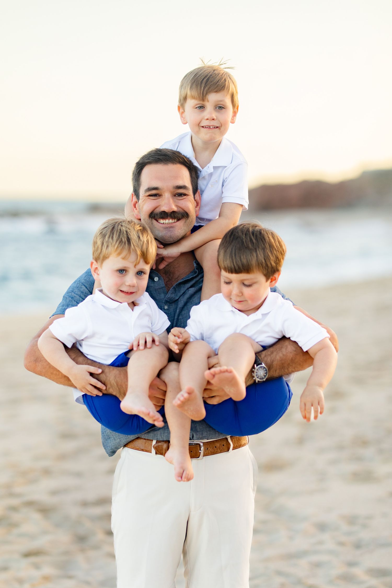 Father smiling on beach holding three young boys, two in arms and one on shoulders, all wearing white shirts.