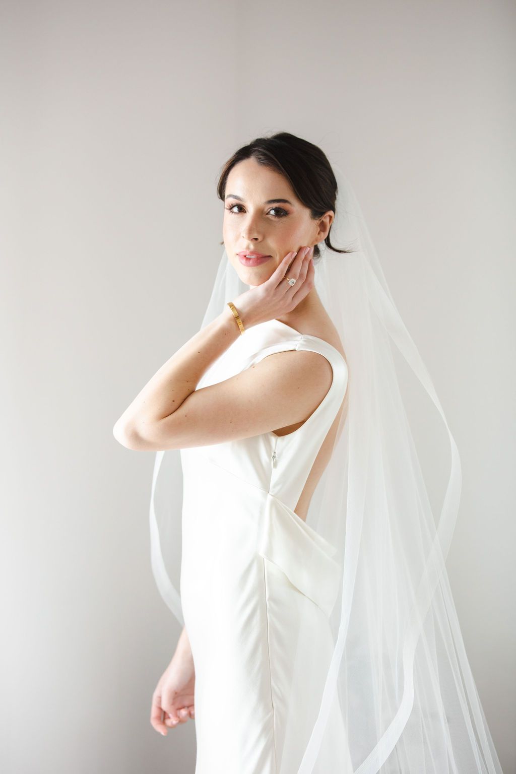 Elegant bride in sleek white gown with flowing veil posing gracefully against white wall.