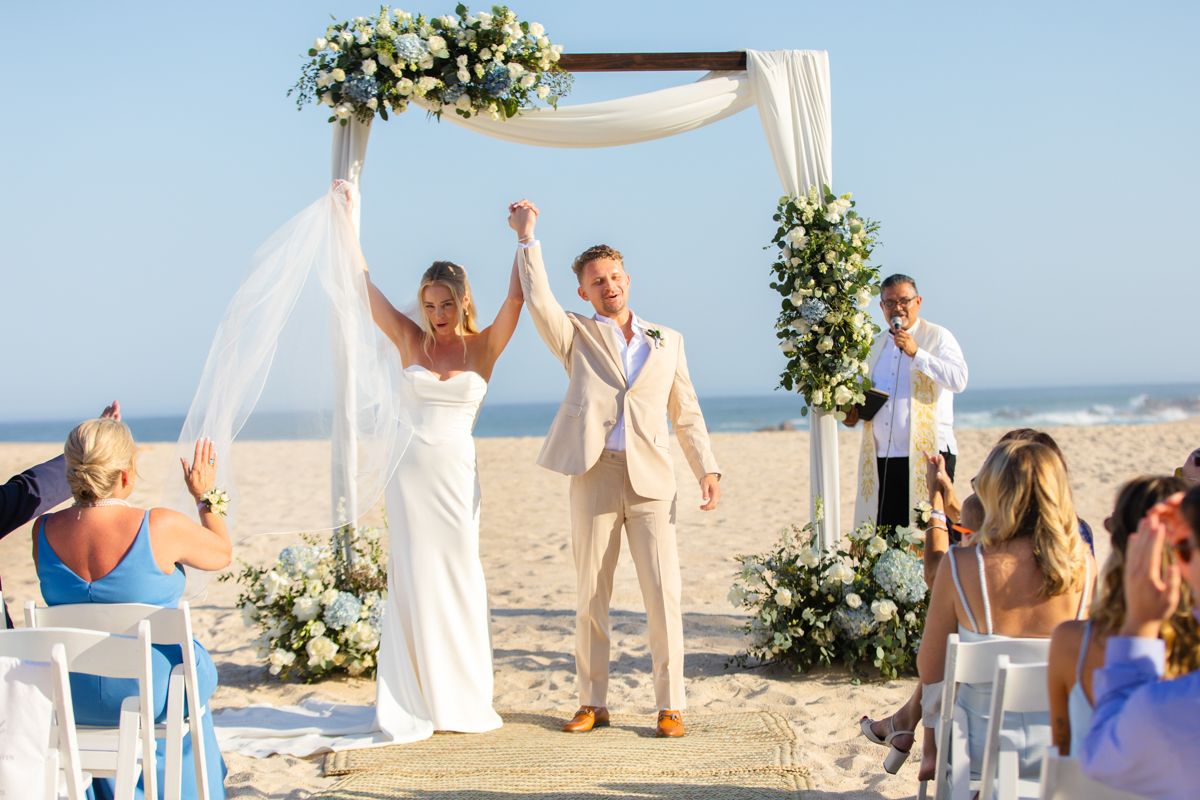 Newlyweds raise hands in celebration at beach wedding altar adorned with white floral arch.