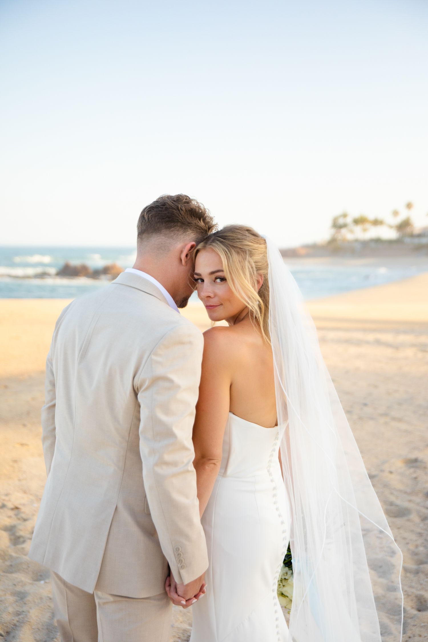 Bride and groom holding hands on a sandy beach, bride in white gown and veil looking back over her shoulder.