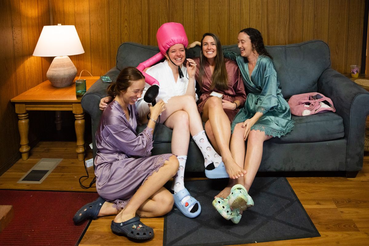 Four bridesmaids laughing together on couch in robes and fun slippers during wedding prep.