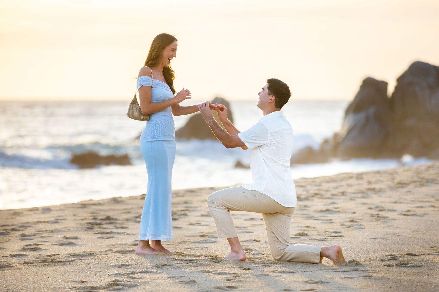 Man proposing to woman on a sandy beach at sunset, kneeling with a ring box as she wears a light blue dress.