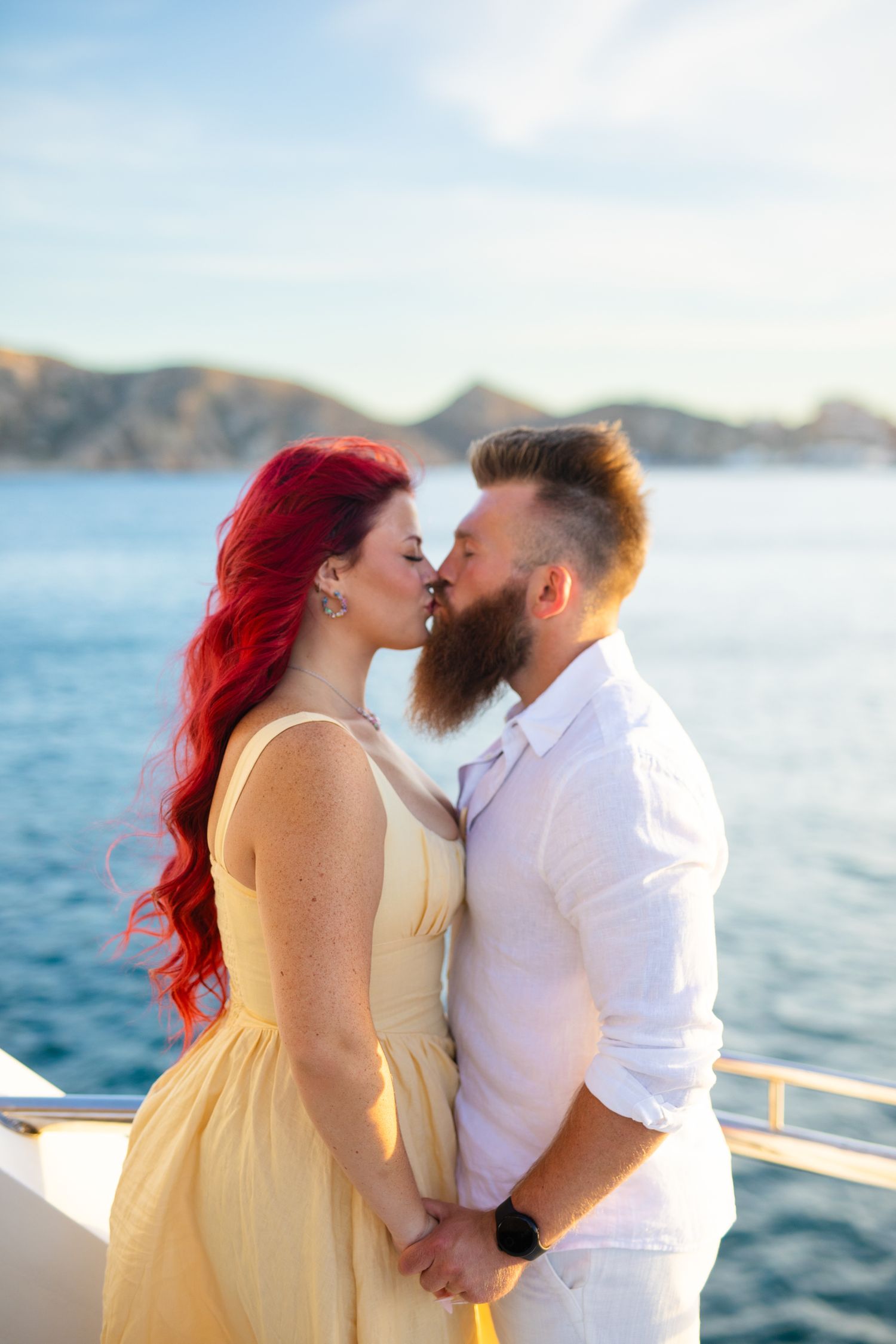Couple kissing on yacht deck, woman with vibrant red hair in yellow dress, bearded man in white, ocean and hills behind.