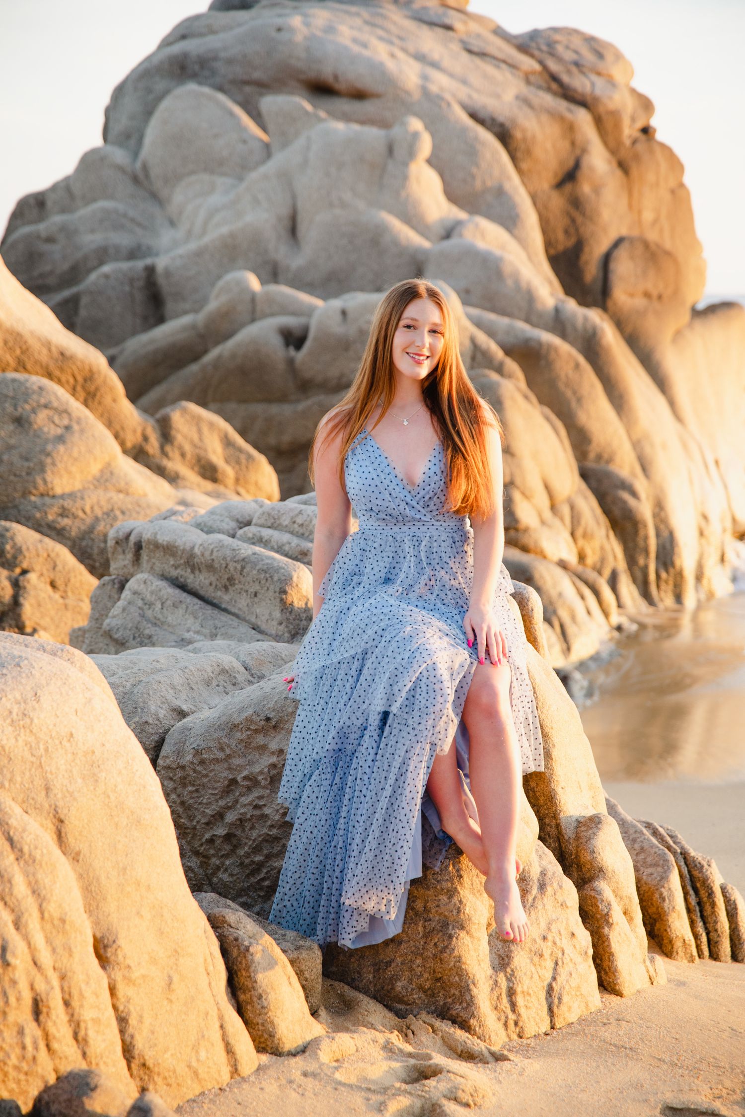 Redhead woman in blue floral maxi dress sitting on rocky boulders at golden hour beach.