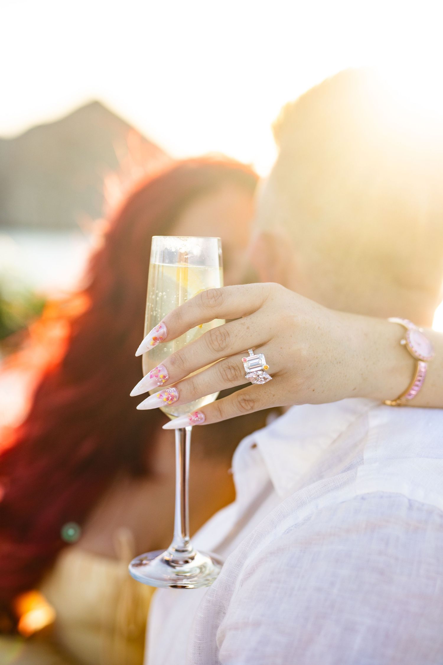 Woman holding champagne flute with large engagement ring and pink floral nails, couple embracing at golden sunset.