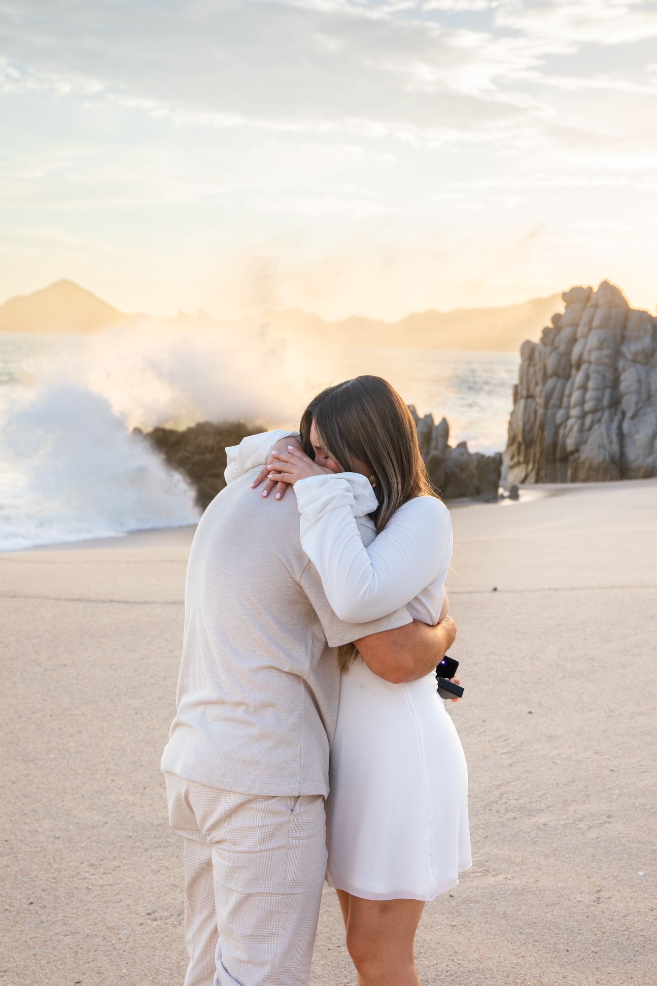 Couple embracing on a sandy beach at sunset with crashing waves and rocky cliffs in the background.