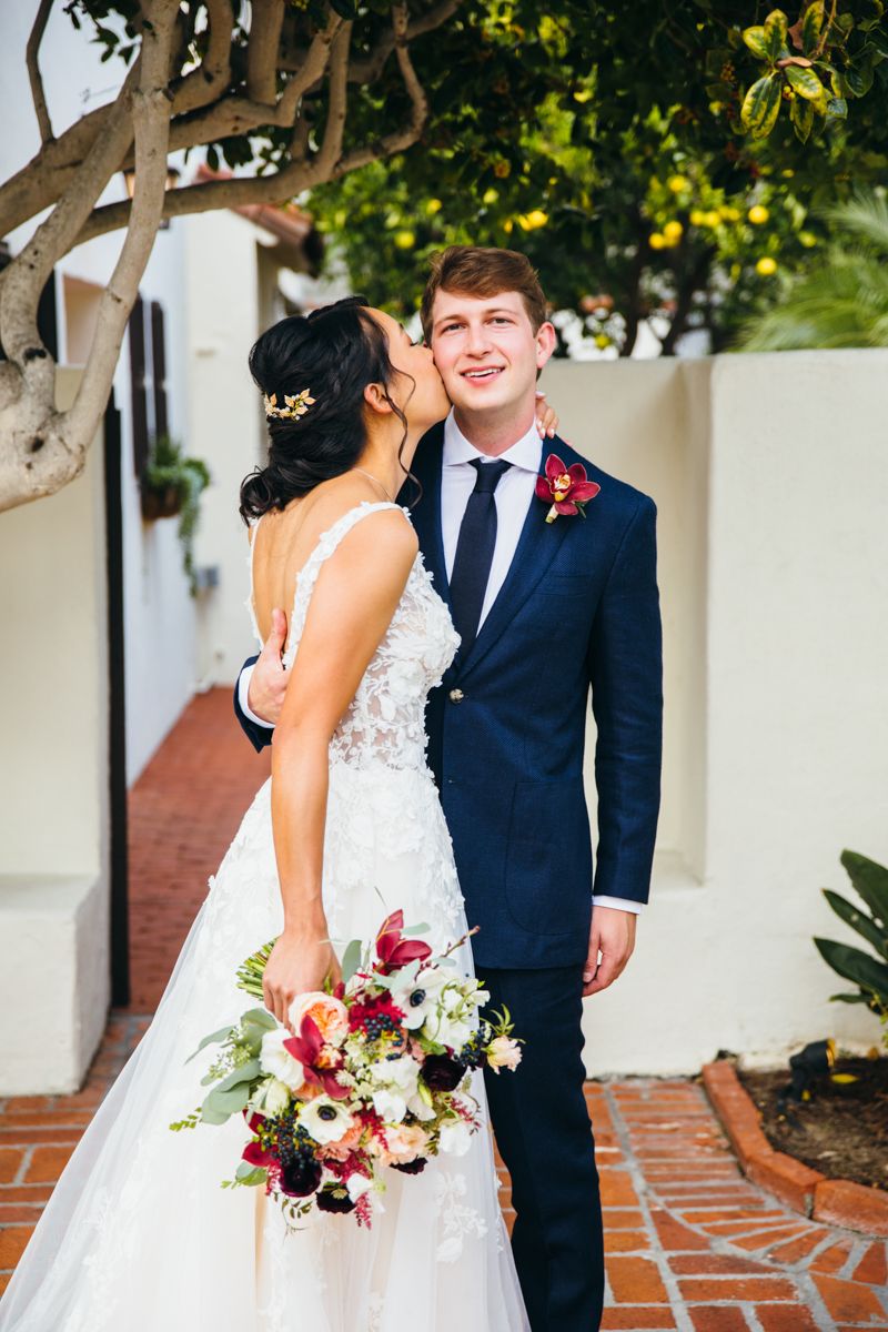 Bride in lace gown kisses groom on cheek while holding a colorful bouquet at an outdoor wedding venue.