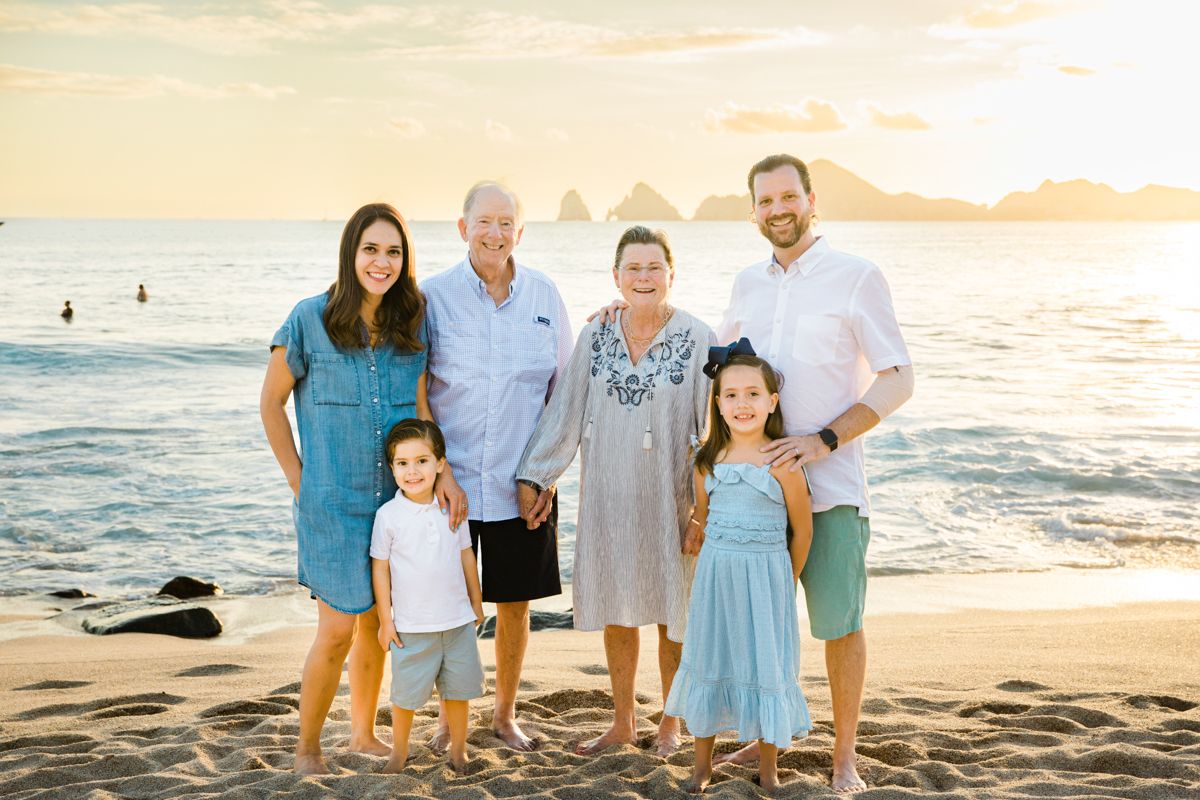 Multi-generational family of six poses together on a sandy beach at golden sunset with rocky coastline backdrop.