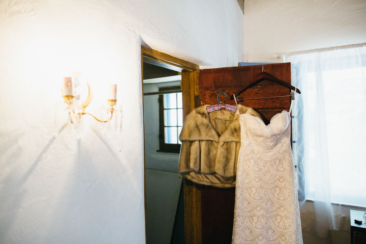A white lace wedding gown and fur stole hang on wooden hangers in a rustic venue doorway.