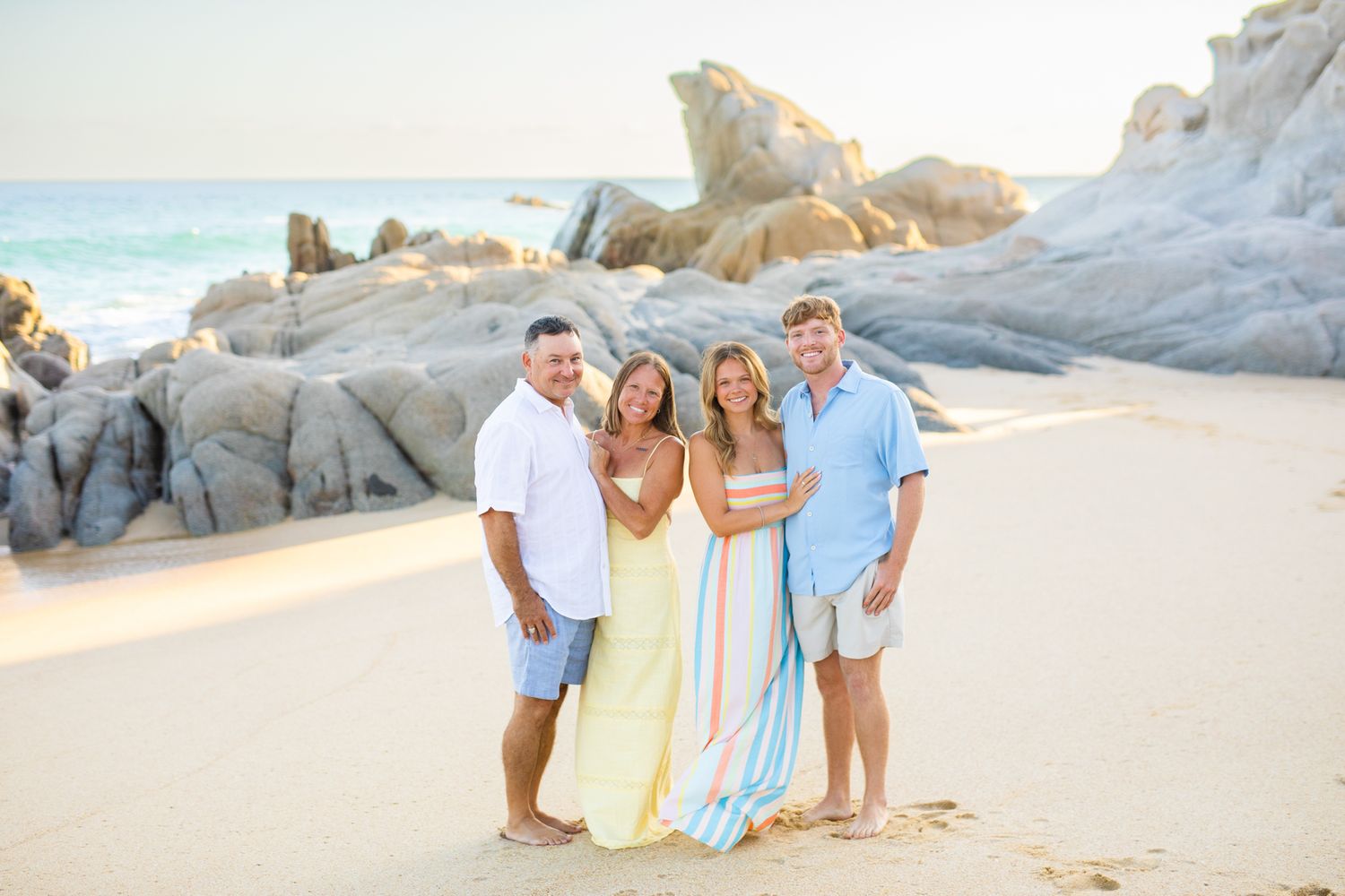 Two couples smile together on a sandy beach with rocky cliffs and turquoise ocean waves in the background in cabo